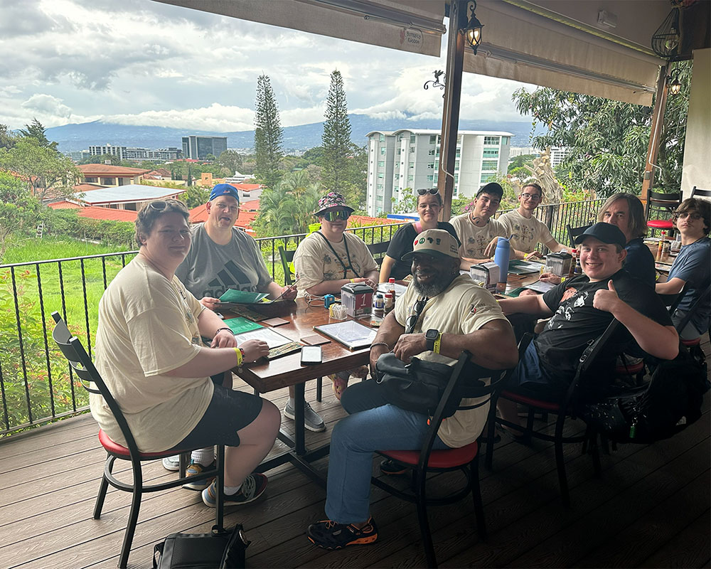 Group of people seated around an outdoor restaurant table on a covered balcony, with drinks and menus visible, overlooking a green hillside and buildings in the distance.