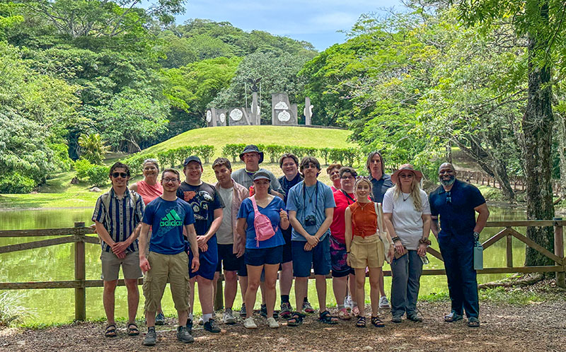 Group of people standing together in front of a pond and wooded hillside, with a monument visible in the background.