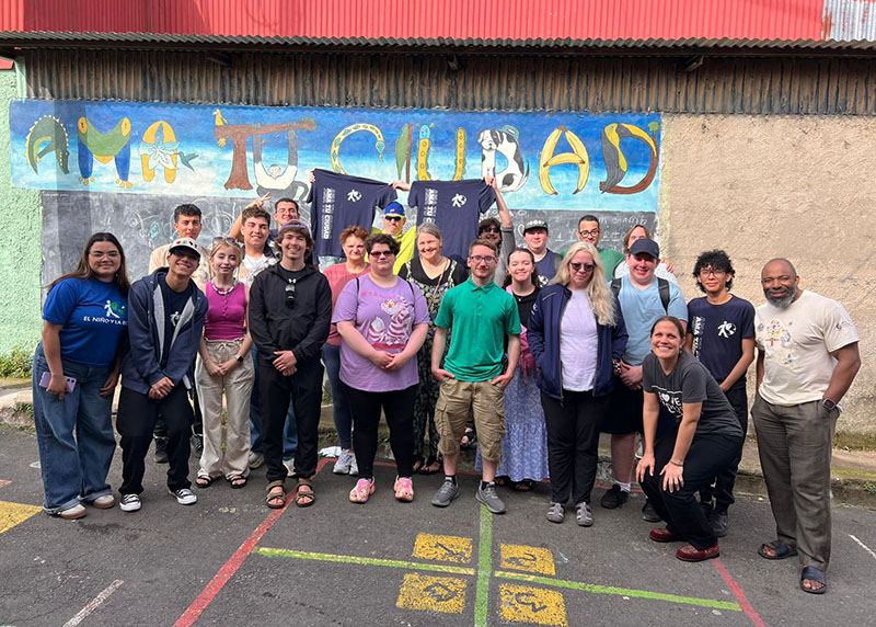 Large group of people posing outdoors in front of a colorful painted wall with the word “Matapalo.” Several individuals hold dark shirts, and the group stands on a paved area marked with painted lines. A corrugated metal roof and textured wall form the background.