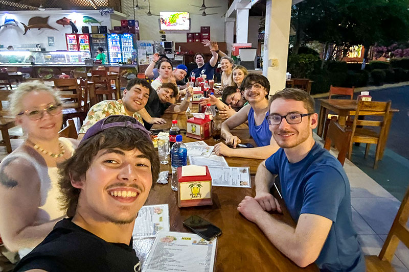 Group of about ten people seated around a long wooden table in an open-air restaurant, taking a selfie together. Menus, drinks, and condiments are on the table, and other tables, chairs, and a lit beverage cooler are visible in the background.