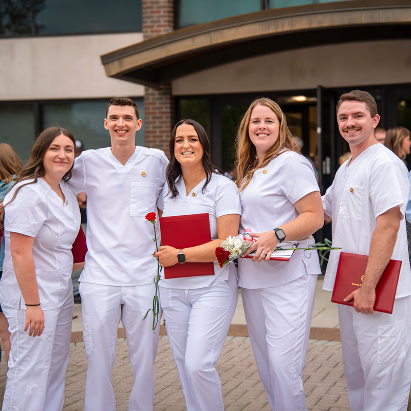 five nursing students holding diplomas and flowers after receiving their nursing pins