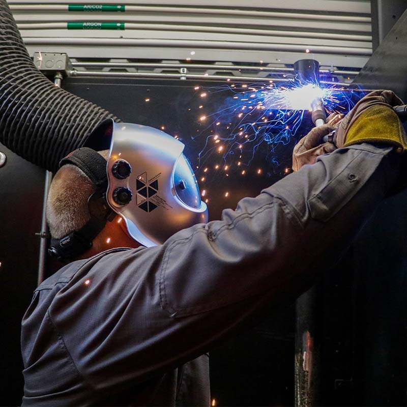 a man in a gray long-sleeved work jacket and protective helmet welding in a workshop bay