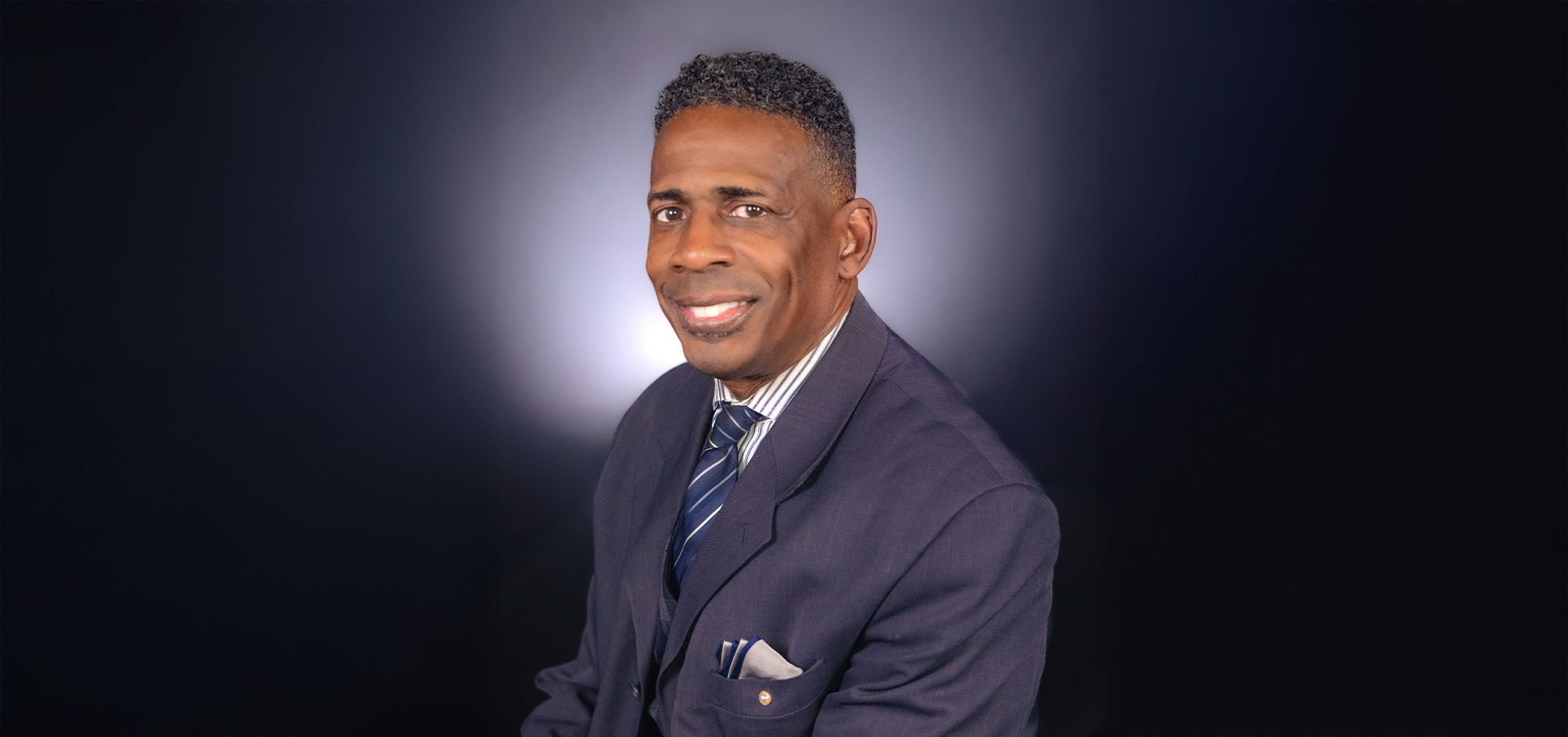 A man in a dark blue suit, with a striped white shirt and striped blue tie, smiling in front of a dark background. 