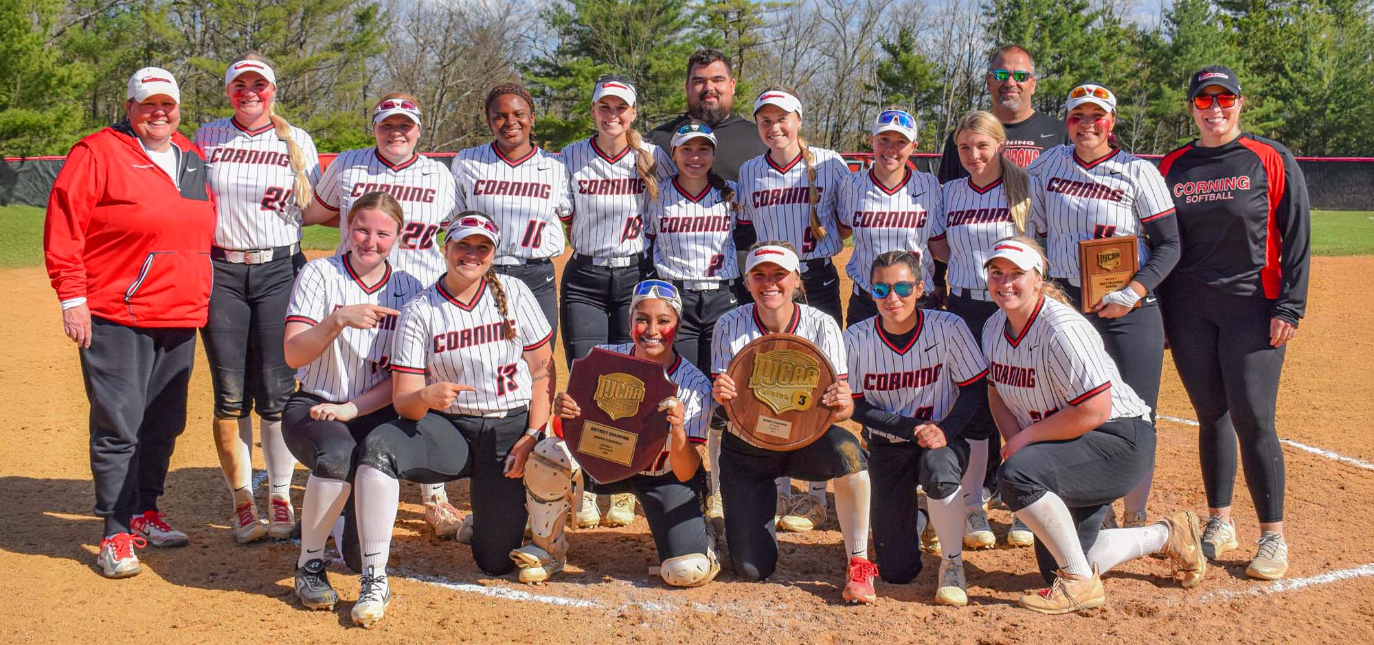 Group of Corning softball players and coaches posing on a softball field, with several athletes kneeling in front holding championship plaques, including a NJCAA Region III softball championship award, while others stand behind in matching white pinstriped uniforms with “Corning” across the front; trees and a clear sky in the background.